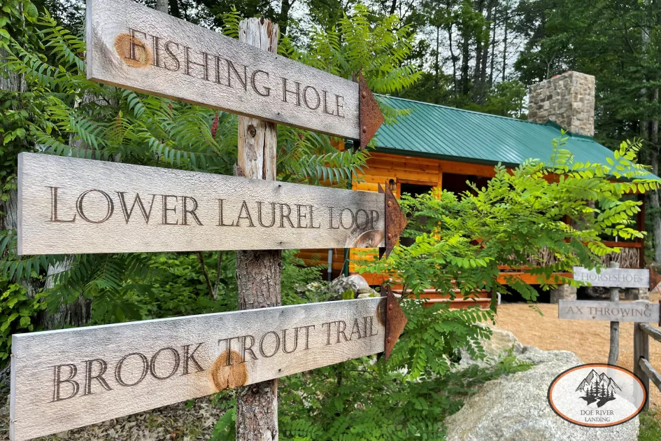 A sign with three plaques points past the Water's Edge Pavilion down a hiking trail. The Sign reads &quot;Fishing Hole,&quot; &quot;Lower Laurel Loop,&quot; and &quot;Brook Trout Trail.&quot;